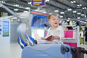 Toddler boy child holding passport with suitcase, sitting on trolley at airport, waiting for departure