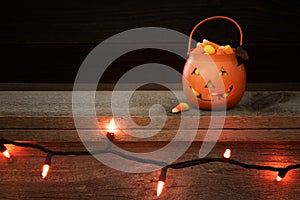 Little Trick or Treat Halloween Pumpkin filled with candy corn on a rustic wood shelf with row of orange lights and dark backgroun