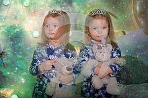 Little twin girls in blue dresses with teddy bears in their hands by the Christmas tree