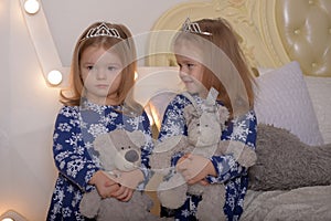 Little twin girls in blue dresses with teddy bears in their hands by the Christmas tree