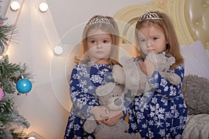 Little twin girls in blue dresses with teddy bears in their hands by the Christmas tree