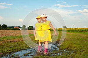Little twin girls in summer yellow dresses and pink boots run through puddles in suburb
