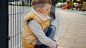 Little upset boy sitting next to metal fence feeling unhappy and lonely. Child depression, problems with bullying, victim in