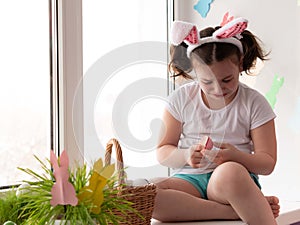 A little upset girl in bunny ears sits alone on the windowsill crying holding a painted shell in her hands, the concept of easter