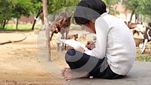 Little village girl doing homework in notebook and sitting on ground near goat farm. Indian countryside footage