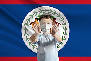 Little white boy in a protective mask on the background of the flag of Belize. Makes a stop sign with his hands, stay at home