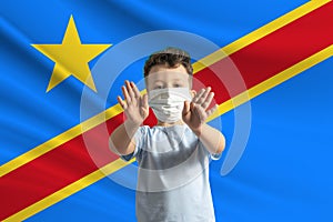 Little white boy in a protective mask on the background of the flag of Democratic Republic of the Congo Makes a stop sign with his