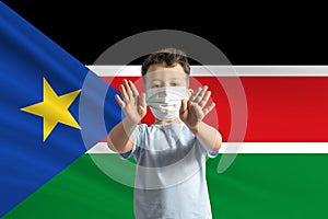 Little white boy in a protective mask on the background of the flag of South Sudan. Makes a stop sign with his hands, stay at home