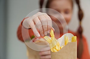 Little white girl eats French fries in a fast food restaurant. Close up photo of a female child taking potato sticks from a box