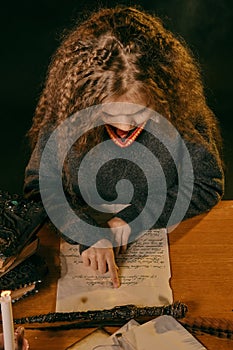 Little witch dressed in dark clothes is sitting at the table against a black smoky studio background and reading a