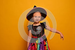 Little witch girl looking at camera and pointing index finger down or holding finger down on yellow background.