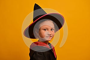 Little wizard girl in Halloween costume on yellow background. Kid in magic hat and mantle or robe looking at camera.