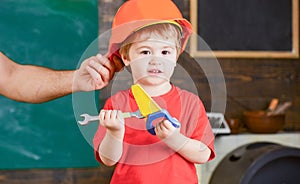 Little worker helping daddy in workshop. Kid playing with toy handsaw and spanner. Father holding protective helmet on