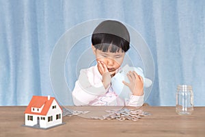 The little yellow-skinned girl holding a piggy bank in hand looks at the coins on the table in a daze