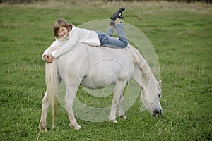 Little young girl in a white sweater and jeans lying on the back of a white pony. Lifestyle portrait