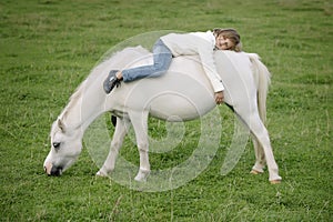 Little young girl in a white sweater and jeans lying backwards on the back of a white horse. Lifestyle portrait