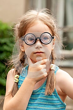 Little young school age girl in big funny quirky glasses thinking, wondering, deep in thought. Outdoors portrait, one person face