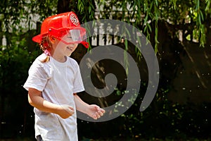 A little young school age girl wearing a red firefighter toy helmet being sprayed with water, outdoors, child playing outside