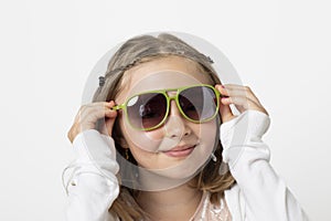 Little young teen girl, posing mischievously in the studio. The background is white and she has really big green sunglasses