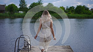 Live camera follows joyful smiling young woman running jumping along wooden pier in slow motion posing. Back view