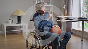 Live camera zoom in desperate Caucasian disabled man in wheelchair with basketball ball. Portrait of hopeless paraplegic