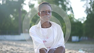 Live camera zoom out as confident smiling African American woman sitting on sandy sunny beach posing. Happy female