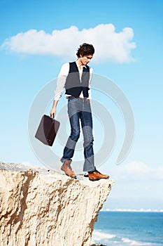 Living on the edge. Young semi-formal businessman stepping towards the edge of a cliff overlooking the ocean.