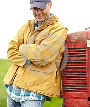Living a good and simple life. a smiling farmer standing next to his tractor with his arms crossed and looking down.