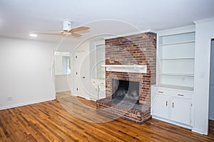 A living room or den with hardwood floors, white walls and a red brick fireplace with mantle in a newly renovated house