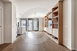 Living room of an empty house with custom-made oak bookcase, open kitchen on one side and terrace with access to a terrace with