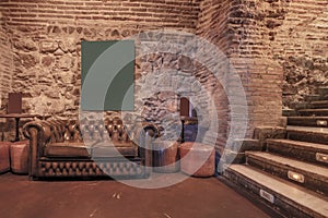 Living room of a nightlife venue with a shiny brown leather chester sofa, exposed stone and brick walls and stairs with indicative