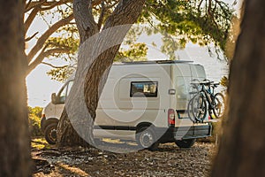 Living the vanlife. White van camper parked on the beach, nice trees around and blue sky just behind the van in the background.