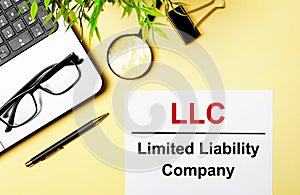 LLC Limited Liability Company is written in red on a white piece of paper on a light yellow background next to a laptop, pen,