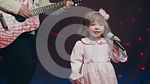 Llittle girl in vintage dress sings on stage, her father plays acoustic guitar