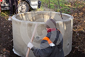 A loader lowers a concrete ring into a dug hole to build a septic tank. A worker installs a sewer into the ground