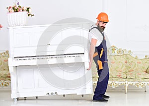 Loader moves piano instrument. Heavy loads concept. Man with beard worker in helmet and overalls lifts up, efforts to