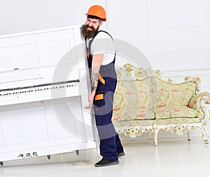 Loader moves piano instrument. Man with beard worker in helmet and overalls lifts up, efforts to move piano, white