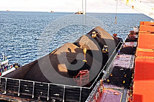 Loading coal from cargo barges onto a bulk carrier using ship cranes and grabs at the port of Muara Pantai, Indonesia. January,202