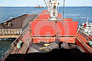 Loading coal from cargo barges onto a bulk carrier using ship cranes and grabs at the port of Muara Pantai, Indonesia. January,202