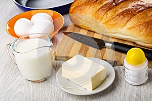 Loaf of bread, knife on cutting board, salt, frying pan, butter in saucer, eggs in bowl, jug of milk on table