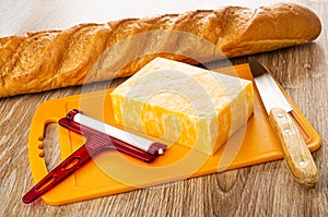 Loaf of bread, piece of marble cheese, cheese cutter, knife on cutting board on wooden table