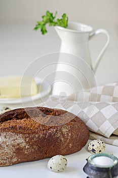 Loaf of rye bread on linen napkin with white jug, salt, quail eggs and butter on background