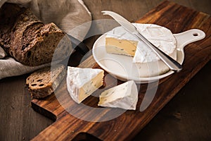 Loaf of soft blue cheese from cow milk on porcelain plate with walnut bread, knife, linen towel and dark brown wooden board as