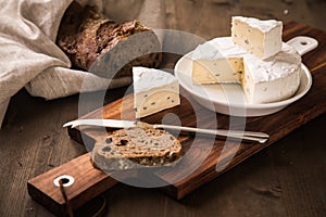 Loaf of soft blue cheese from cow milk on porcelain plate with walnut bread, knife, linen towel and dark brown wooden board as