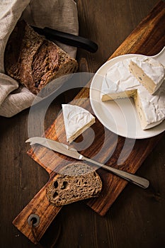 Loaf of soft blue cheese from cow milk on porcelain plate with walnut bread, knife, linen towel and dark brown wooden board as