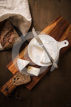 Loaf of soft blue cheese from cow milk on porcelain plate with walnut bread, knife, linen towel and dark brown wooden board as