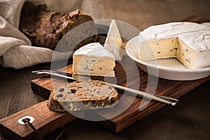 Loaf of soft blue cheese from cow milk on porcelain plate with walnut bread, knife, linen towel and dark brown wooden board as