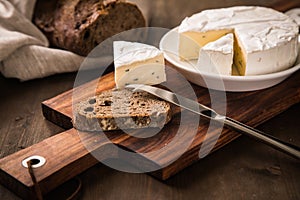 Loaf of soft blue cheese from cow milk on porcelain plate with walnut bread, knife, linen towel and dark brown wooden board as