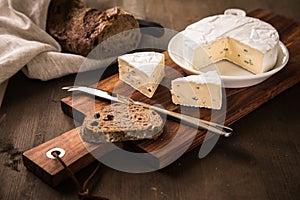 Loaf of soft blue cheese from cow milk on porcelain plate with walnut bread, knife, linen towel and dark brown wooden board as