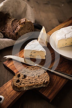 Loaf of soft blue cheese from cow milk on porcelain plate with walnut bread, knife, linen towel and dark brown wooden board as